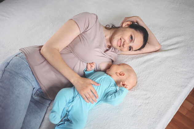 分娩年轻的母亲躺在床上 抱着一个穿着蓝色连体衣的可爱的新生婴儿 用母乳给他喂奶母乳喂养可爱爱