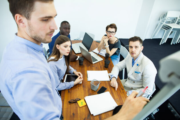 演示讨论公司进步自信年轻人站在白板旁边 指着图表 而他的同事坐在办公桌旁团队室内场所
