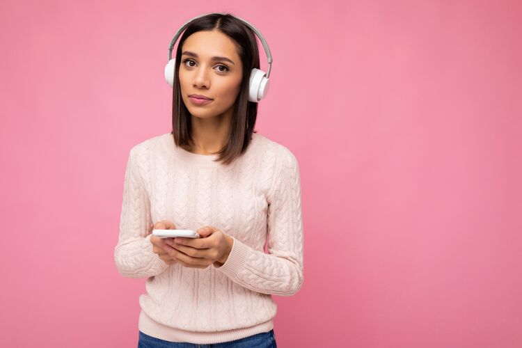 休闲漂亮可爱的年轻女子穿着粉色毛衣隔着粉色的墙穿白色音频电话无线