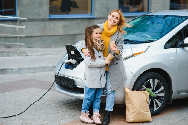 女性妈妈带着女儿在电动加油站给电动车充电 用手机说话充电移动性白天