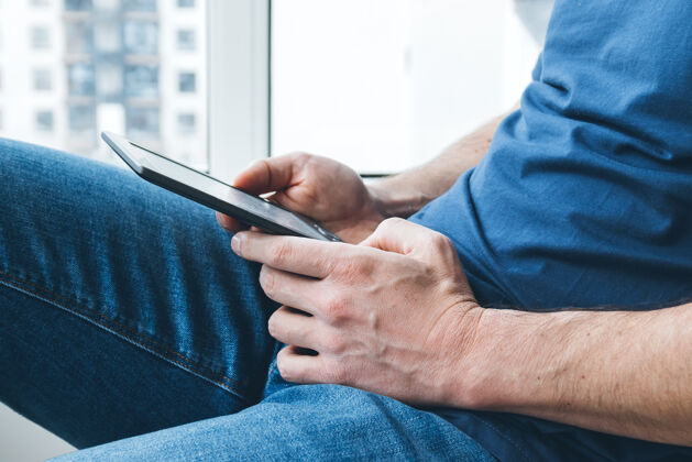 举行一名身穿蓝色t恤和牛仔裤的男子手里拿着一本电子阅读器电子书手阅读家里的书窗口选择集中网络在线电子