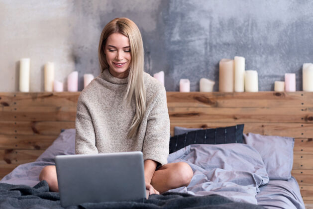 卧室休息回家很高兴美丽的年轻女子坐在床上 一边用笔记本电脑一边放松毛衣享受蜡烛