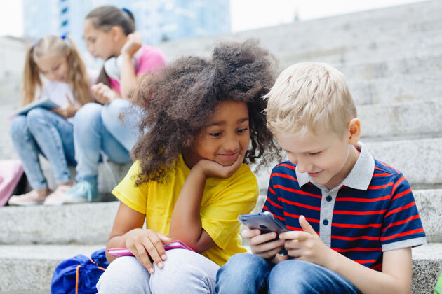 学校两个小学生：一个非洲裔美国女孩和一个金发男孩正在看一部智能手机 在一个阳光明媚的夏日户外坐在台阶上活动欢呼人