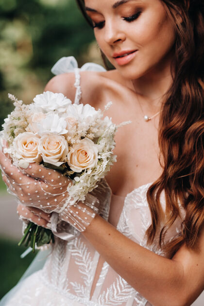 手套一个优雅的新娘在一个自然公园里穿着白色的裙子和一束花束的肖像时尚年轻美丽
