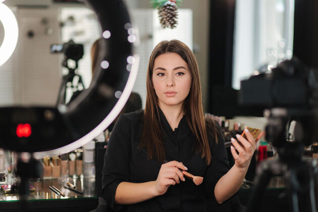 年轻人年轻漂亮的女人和专业的化妆师博客记录化妆教程在工作室摄影棚拍摄视频博客模特