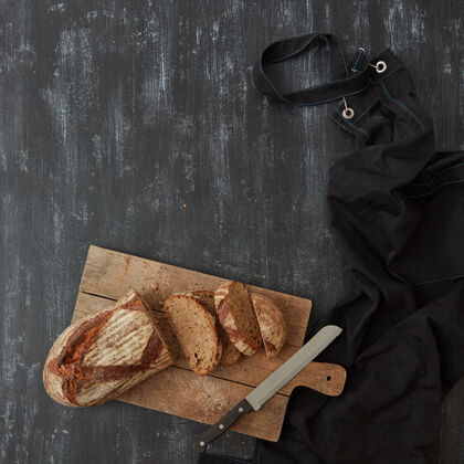 浅一块土生土长的面包 放在砧板上 用刀和黑色围裙平铺在深色的背景上面包房切片种子