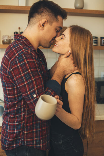 男人早上 一对美丽迷人的情侣在厨房里亲吻 而男人正抚摸着他女朋友的脖子 而她正端着一杯茶浪漫亲吻男朋友