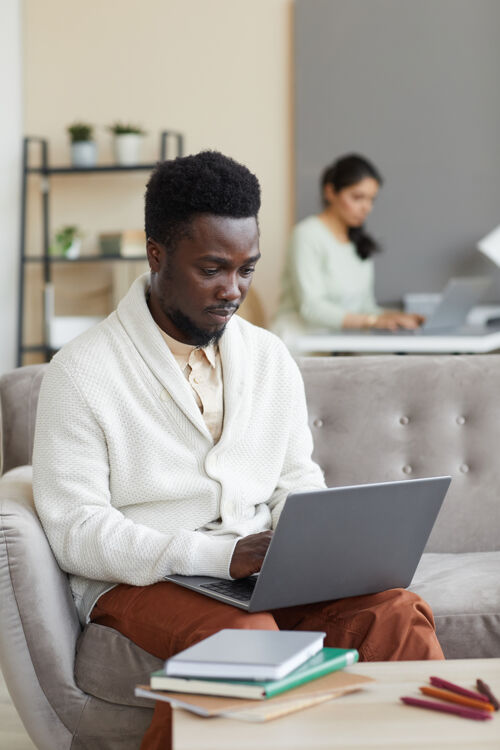 快乐一个非洲男人坐在桌子前的沙发上 手里拿着书 在笔记本电脑上打字 他在家里在线学习笔记本电脑互联网专业职业
