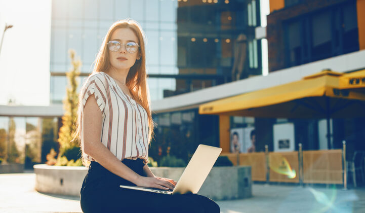 行政一位迷人的红发女学生 课后坐在室外一栋楼附近 腿上放着一台笔记本电脑 看着夕阳笔记本电脑眼镜城市