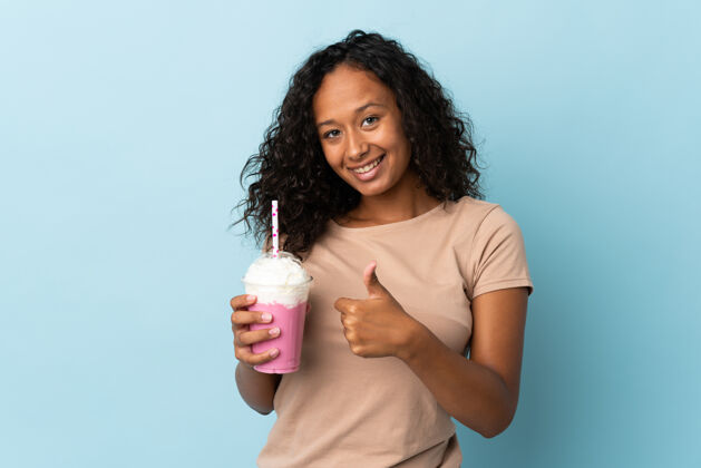 女人一个十几岁的女孩拿着草莓奶昔孤立地站在蓝色背景上竖起大拇指头发牛奶冰沙