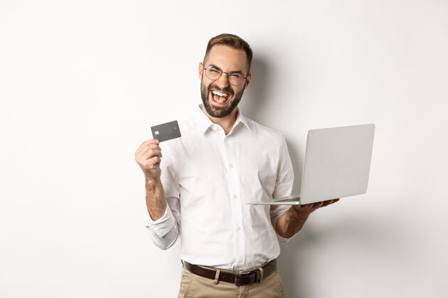 Laptop在线购物帅哥男子出示信用卡 用笔记本电脑在网上订购 站在白色背景上Professional买家房地产经纪人
