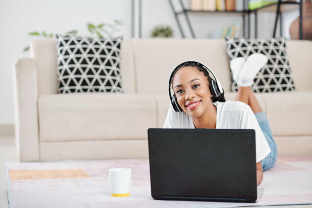 编程正面的年轻黑人妇女戴着耳机躺在地板上 在家里用笔记本电脑工作的肖像家庭电脑微笑
