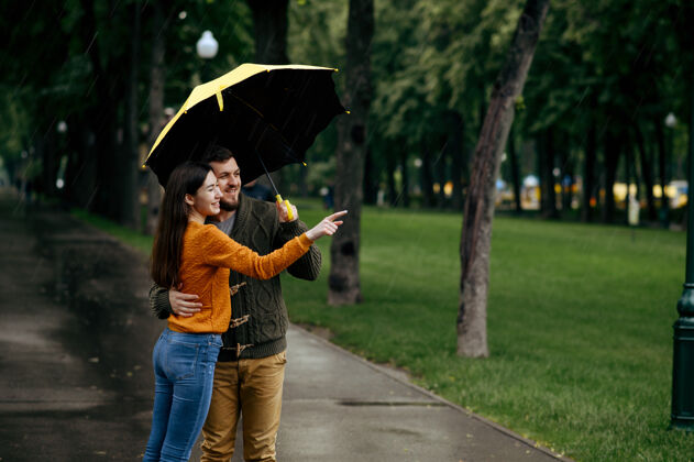 湿快乐的情侣在公园里悠闲 夏日多雨戴曼雨中的女人站在雨伞下 漫步小径上的浪漫约会 小巷里的潮湿天气丈夫保护男人