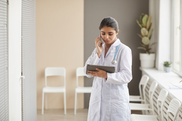 医院穿着白大褂的年轻女医生站在医院的走廊上用手机和数字平板电脑上网医药职业室内