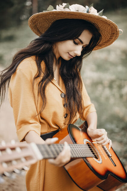 音乐一位留着深色长发的美丽女子的肖像 戴着帽子 在大自然外面的吉他上唱歌成人音乐家经典