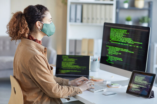 人员戴着防护面具的年轻开发人员在办公室里看着电脑显示器 在电脑键盘上打字通信职业流行病