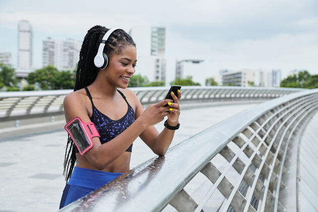 聆听微笑迷人的年轻混血女子站在桥上 通过智能手机上的应用程序查看她的跑步轨迹慢跑女性选择