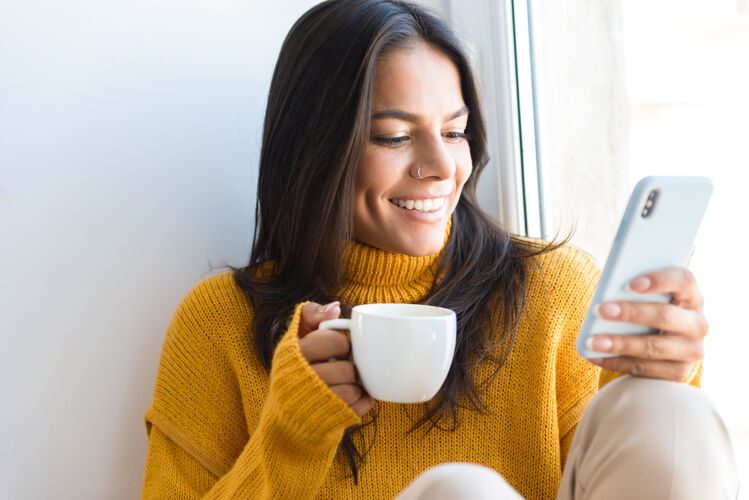咖啡特写镜头中 一位穿着毛衣的可爱年轻女子坐在室内的窗前 端着一杯茶 用手机欢呼饮料成人