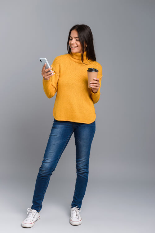 白种人一个随意的年轻女子的全长肖像 隔着灰色的墙 拿着外卖咖啡用手机应用程序微笑女性
