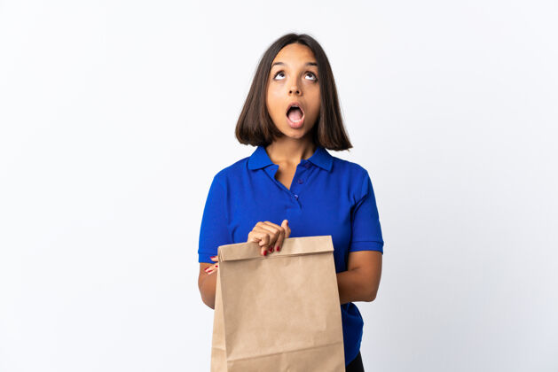 表情一个年轻的拉丁女人拿着一个杂货店的购物袋 孤零零地站在白色的地板上 抬起头来 脸上露出惊讶的表情幸运年轻顾客