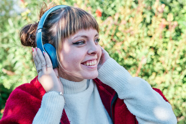年轻人一个穿着红色夹克 戴着蓝色耳机在公园里听音乐的白人女孩时尚模特耳机