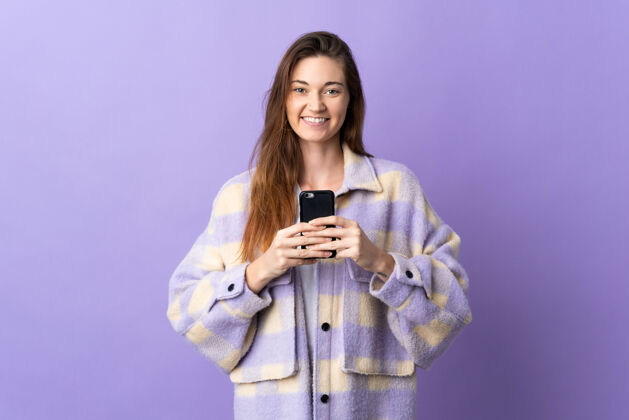 女孩年轻的爱尔兰女子在紫色背景下看着相机 微笑着使用手机电话漂亮女性