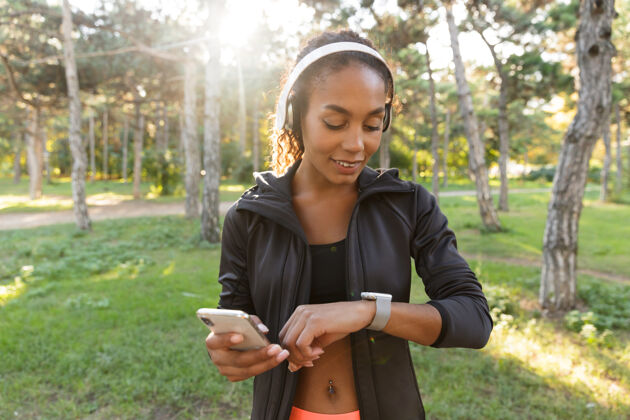 女士一幅20多岁的快乐女人的画像 穿着黑色运动服 戴着耳机 一边看着手表 一边穿过绿色公园运动年轻成人