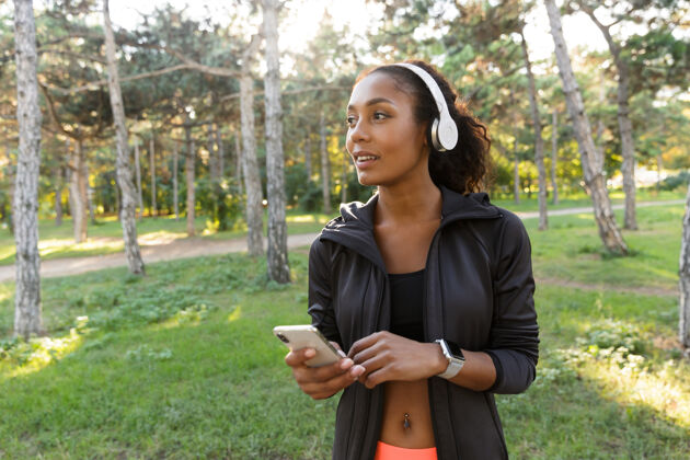 精力充沛20多岁的黑发女子穿着黑色运动服 戴着耳机 一边在绿色公园里散步一边用手机移动训练女性