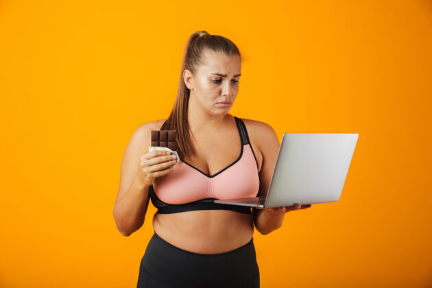 悲伤一个穿着运动服的失望的超重年轻女子的肖像 孤立地站在黄色的墙上 用笔记本电脑 拿着巧克力棒笔记本电脑健身电脑