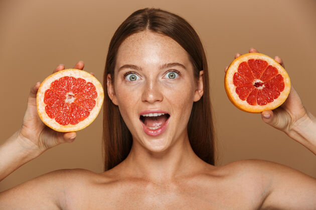 时尚美丽的肖像 一个美丽健康的年轻裸体女子长着红色的头发显示切片柚子隔离在米色墙上一半脸搞笑