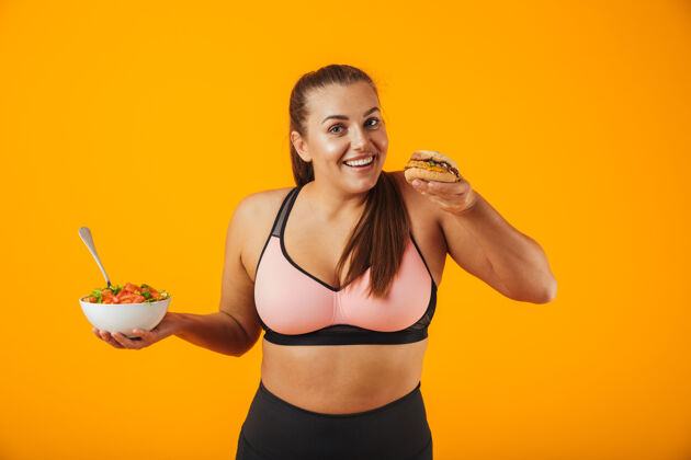 训练一个穿着运动服的快乐的超重健身妇女的肖像 孤立地站在黄色的墙上 手里拿着盛着沙拉和汉堡的碗吸引力蔬菜午餐