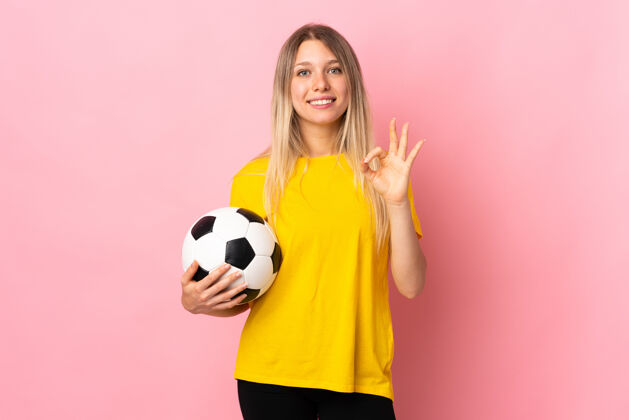运动装一个年轻的足球运动员 一个被隔离在粉红色皮肤上的女人 用手指表示ok专业女人球