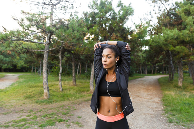 耳机一幅20多岁严肃的非洲裔美国妇女的画像 她穿着黑色运动服 在绿色公园里锻炼身体女性温暖训练