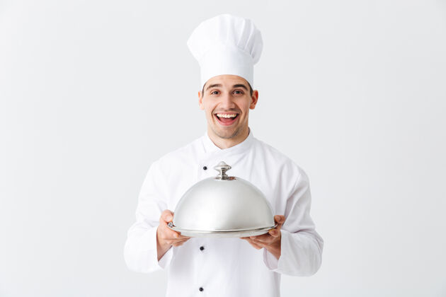 厨师兴奋的男人厨师穿着制服拿着cloche隔着白墙帽子食物概念