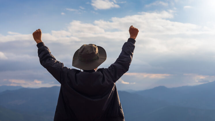 男性快乐的旅行家站在山顶举手致意游客云风景
