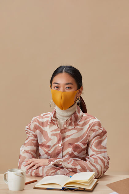 成人戴着防护面具的亚洲妇女坐在桌子旁学习时注视前方的画像商务人士学生书