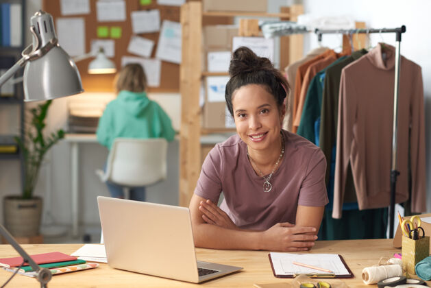 企业家时装设计师坐在工作间笔记本电脑前的桌子前微笑的画像裁缝工作时尚