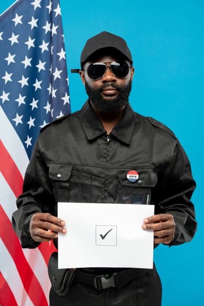 符号当代年轻的非裔美国保安人员身穿黑色制服 戴着墨镜 拿着方格上打勾的选票 对着美国国旗军队美国国旗控股