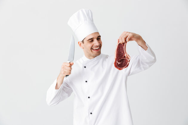 烹饪开朗的厨师穿着制服展示生牛排拿着刀隔着白墙烧烤菜烤架