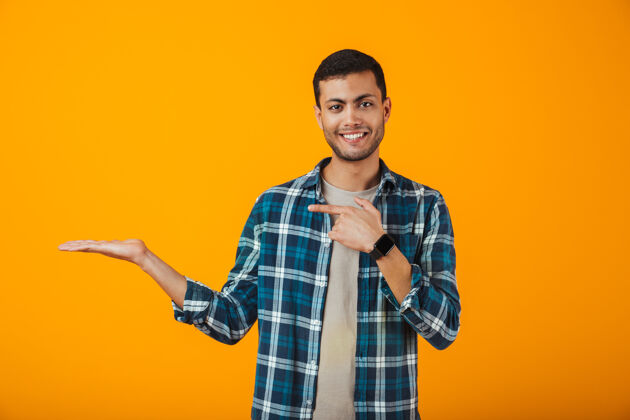 指向一个穿着格子衬衫的快乐的年轻人孤零零地站在橙色的墙上 在他的手掌上展示着复制的空间方格手掌男性