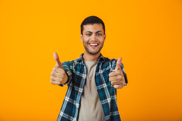 看一个穿着格子衬衫的快乐的年轻人孤立地站在橙色的墙上 竖起大拇指自信肖像白种人