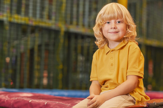幼儿公园里坐在蹦床上金发小男孩的画像特写快乐肖像
