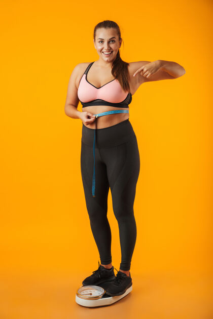 减肥一个穿着运动服的满意的超重年轻女子站在隔离在黄色墙壁上的天平上 腰间拿着卷尺的全长肖像肥胖纤维尺寸