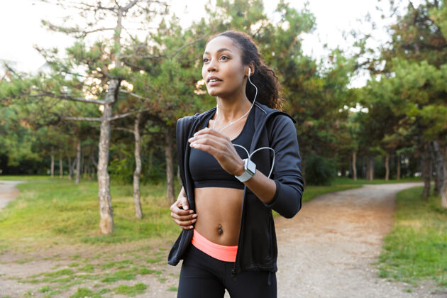 吸引力20多岁的快乐女人穿着黑色运动服 戴着耳机 在绿色公园里跑步自然健身女孩