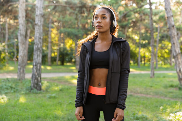 散步20多岁的年轻女子穿着黑色运动服 戴着耳机 穿过绿色公园吸引力精力充沛成人