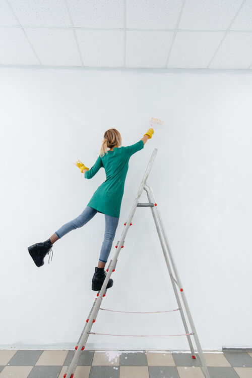 公寓一个年轻的女孩在梯子上用滚筒粉刷白色的墙壁修理内部室内家庭颜色