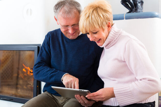 丈夫生活质量——两位老人坐在炉子前的家里 在平板电脑上写电子邮件妻子阅读成熟