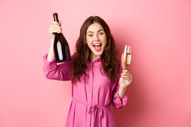 快乐庆祝和节日的概念快乐的女人跳舞 玩得开心 喝香槟 拿着酒瓶和玻璃杯 烤面包 站在粉红色的墙上惊人情绪派对
