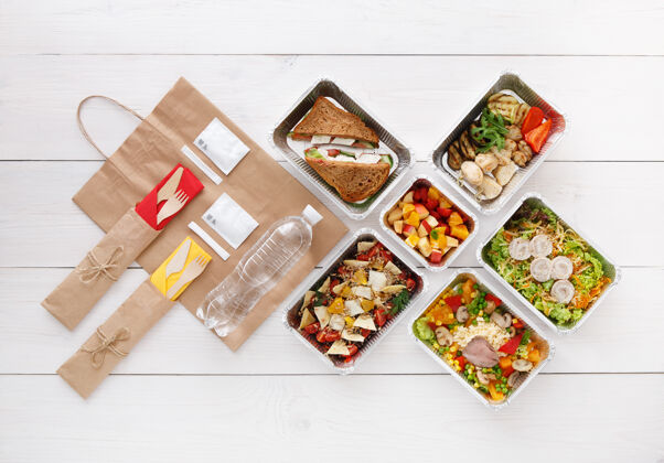 蔬菜健康食品配送外卖食品铝箔盒中的蔬菜 肉类和水果 餐具 水和牛皮纸包装俯视图 平放在白色木头上 有复印空间包装纸烹饪