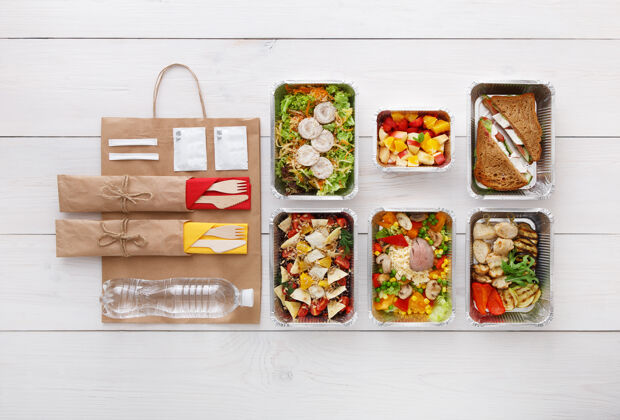 美食健康食品配送外卖食品铝箔盒中的蔬菜 肉类和水果 餐具 水和牛皮纸包装俯视图 平放在白色木头上 有复印空间平铺顶视图美食